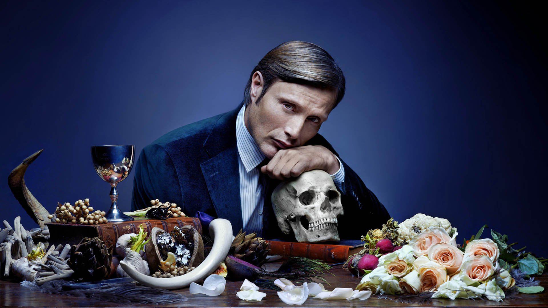 مدس میکلسن پشت یک میز در سریال Hannibal و یک جمجمه در دست او